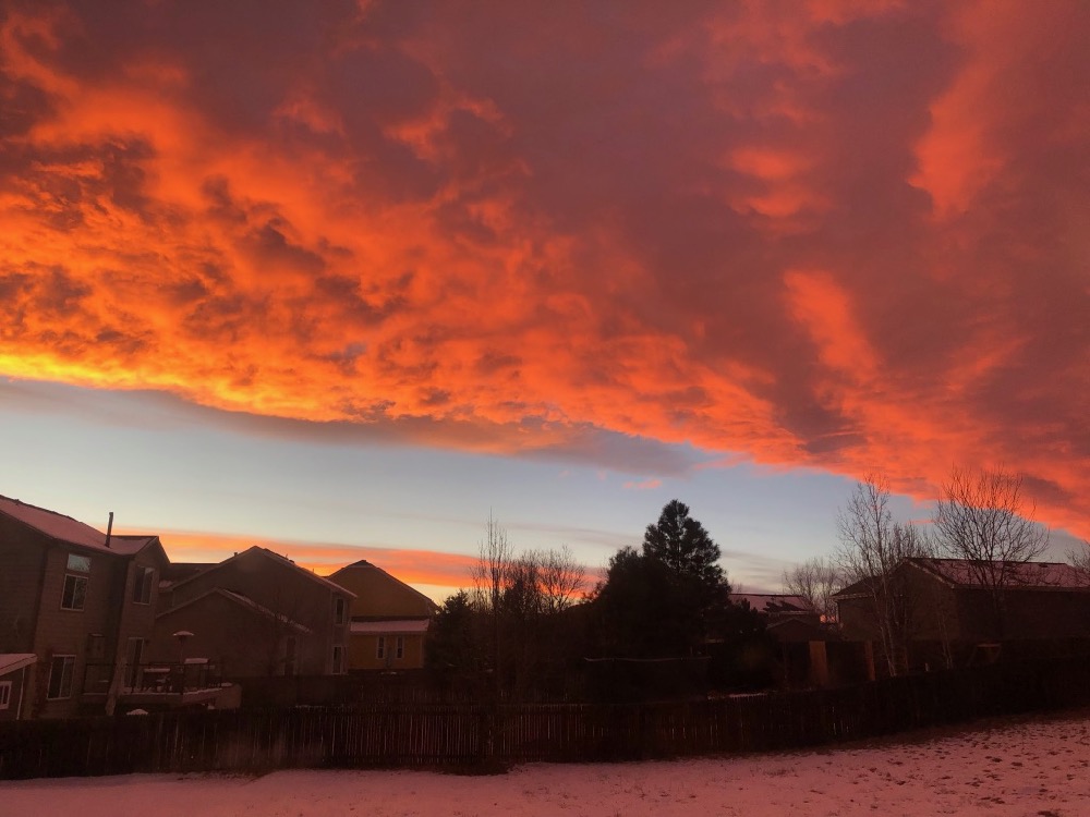 Yellow, Pink, and Orange clouds with some blue sky over houses, trees, and snow.