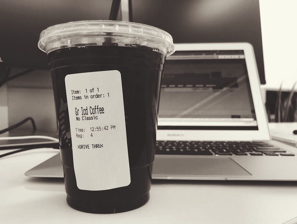 A clear plastic cup of black coffee with ice in it, positioned in front of an open laptop and monitors.