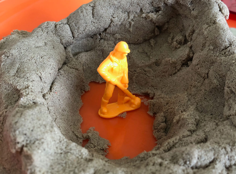 Plastic orange army man standing in a hole amidst kinetic sand