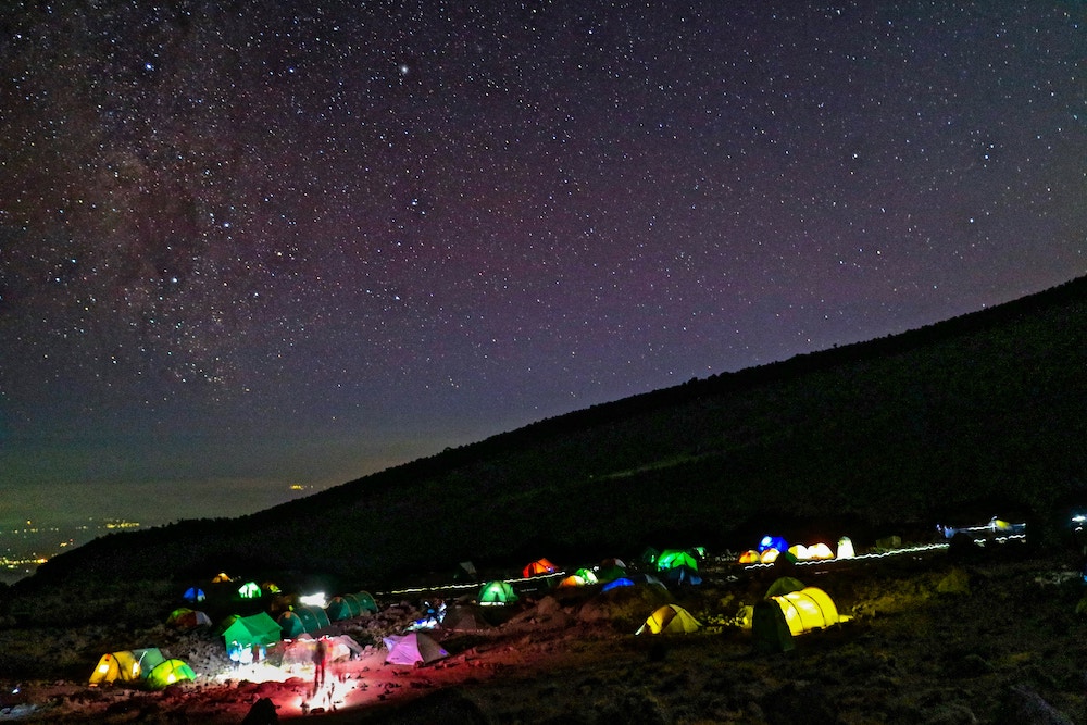 Night sky with clearly visible stars and a campsite below with many tents lit internally.